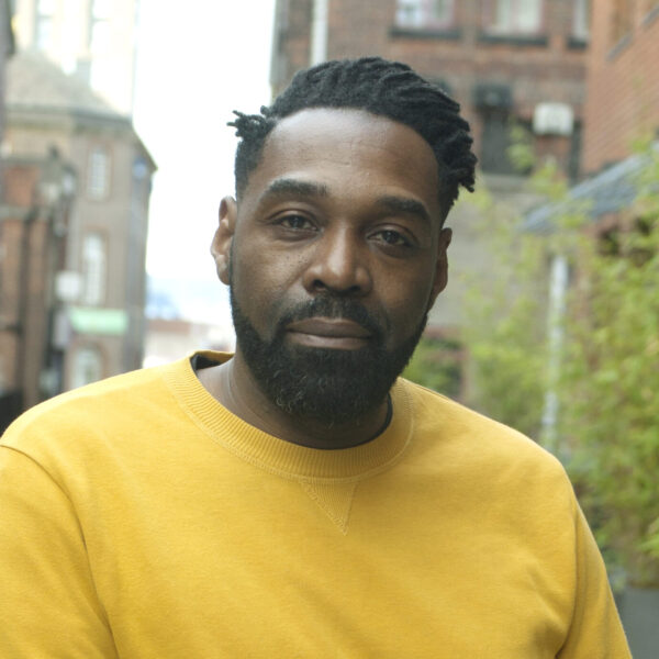 A man with a beard wearing a yellow shirt poses in an alley, flanked by rustic brick walls and dim lighting.