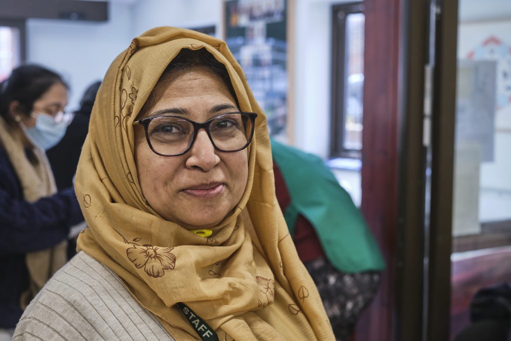 In a bright room, a woman wearing a headscarf and glasses stands, showcasing her unique style and personality.